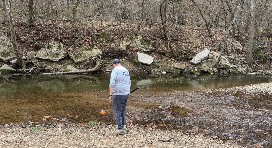 Man standing by a stream with rocks and trees in the background retrieving disc from water with Max Disctance suction cup ball retriever